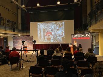 students seated in the 900 room, looking toward the front at Tom Marshburn on a screen in a Davidson t-shirt in a spaceship with two in-person people behind podiums on both sides of the screen