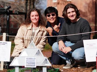 three students smiling with a tiny house in front of them