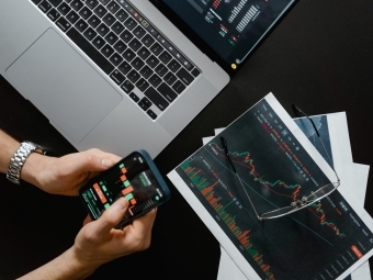 Handings holding phone overlooking laptop and stock projections on piece of paper