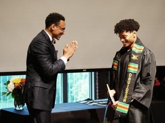 Two Black men smile as one puts graduation regalia around himself