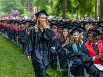 Female student walks in graduation regalia and holds a hand to her face 