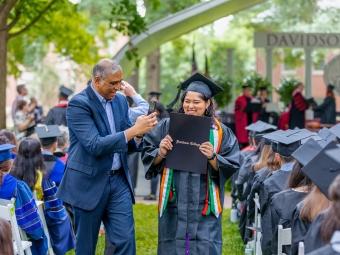 Student holds diploma and smiles while father takes picture
