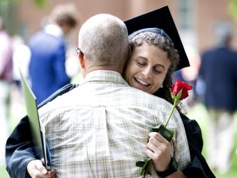 Female student in cap and gown embraces her father while holding a flower