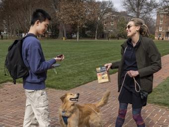 A white woman with blonde hair hands a granola bar to a student while holding a dog