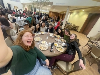 A group of students take a selfie together while sitting around a round dining table in a dining hall