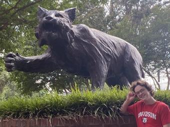 a young student wears a Davidson t-shirt while smiling and standing in front of a wildcat statue