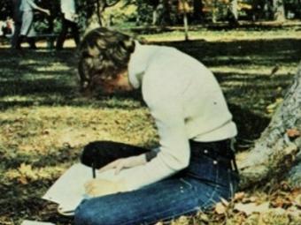 a young man sits wearing jeans and a sweater under a tree while working on a paper