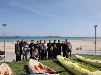 a group of students in wetsuits standing in front of a beach with their paddleboards