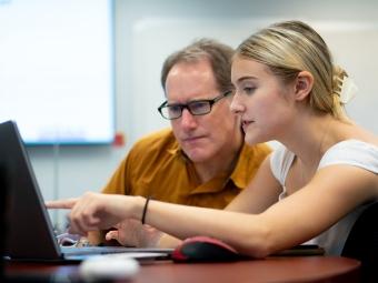 a young woman and an older man huddle around a laptop together while talking