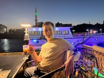 a young man sits on a patio along a river while holding a coffee and smiling