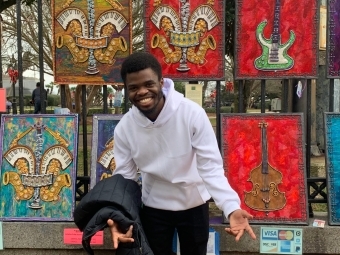 a young Black man wearing a white sweatshirt stands in front of a wall of paintings of musical instruments