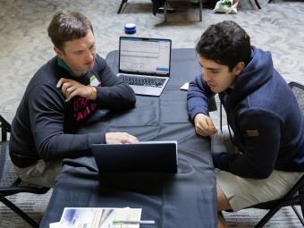a young man talks to an older man while looking at a laptop screen