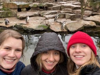 three young women stand in front of a zoo filled with penguins