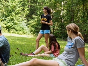 three young people stand and sit in a grassy field with woods in the background