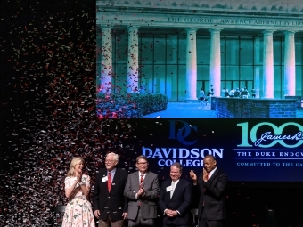 a group of five people standing on a stage clapping as confetti falls around them