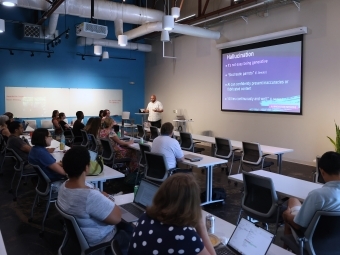 a modern classroom where adults sit around on laptops listening to a speaker
