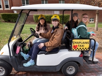 a group of four students sit in a golf cart with a sign that reads "Chaplains' Tax"