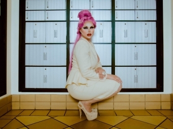 a young white woman in drag crouches in front of a wall of post office boxes