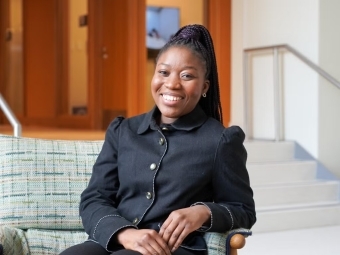 a young Black woman wearing a black jacket sits at a couch smiling