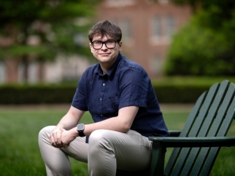 a young white man with a collared blue shirt sitting on a green adirondack chair on a college campus