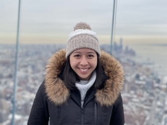 a young woman stands on the roof of a skyscraper in New York City, she wears a beanie and big coat and the sky appears to be grey and cloudy