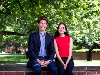 a young man and a young woman sit together on a brick wall in front of green trees