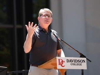 an older white man speaks at a lectern with "Davidson College" on it on a sunny day outside