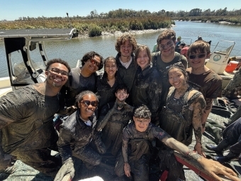 a group of young people stand together, covered in mud, along a riverbank on a sunny day