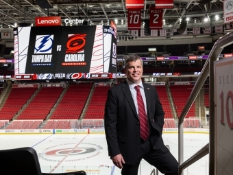 a middle aged white man wearing a black suit with a red tie in front of the Carolina Hurricanes hockey rink