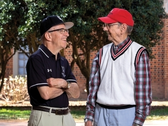 two older men stand together smiling looking at each other in front of a stadium, wearing Davidson College gear