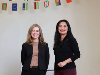 two women standing together in front of a white wall with international flags hanging in the background