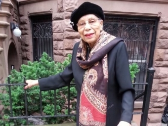 a Black woman wearing a cap and scarf standing in front of a brownstone