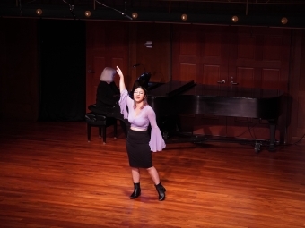 a young woman sings on stage in a purple blouse and black skirt