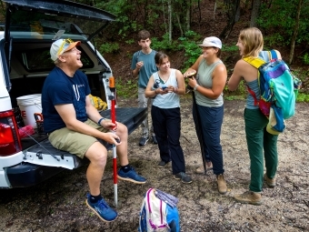 a group of students with their professor gathered around a truck in the woods wearing hiking gear
