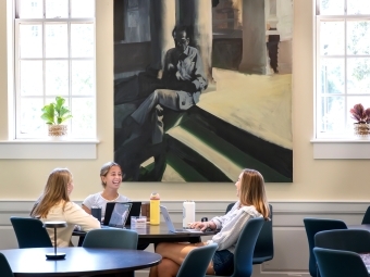 three young women smile while gathered around a table with laptops and waterbottles