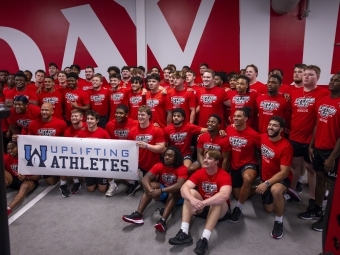 Male student athletes smile in a group behind an event banner with purple text