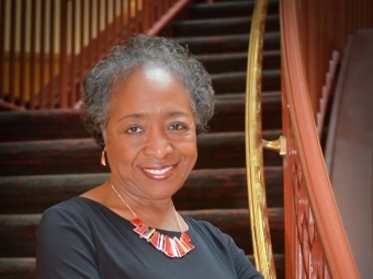 Adult woman in black dress smiles with hands folded in front of grand staircase