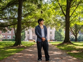a young man stands on a college campus looking to his side in front of a large building surrounded by trees