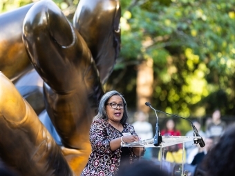 a Black woman speaks at a lectern in front of a sculpture of hands