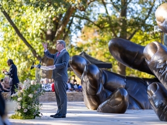 President Doug Hicks at the With These Hands dedication, addressing the crowd at a podium in front of the sculpture