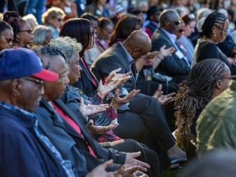 A group of seated guests at the With These Hands sculpture dedication, holding their hands up in the air to emulate the sculpture