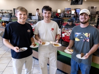 Three smiling male students hold two plates of pie each