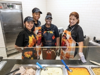 Four female Commons employees wear turkey aprons, hair nets/hats, and gloves behind large serving dishes of macaroni and cheese and mashed potatoes