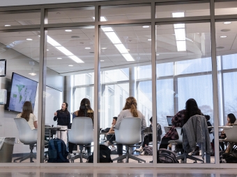a professor stands in a small glass-encased classroom pointing at a screen