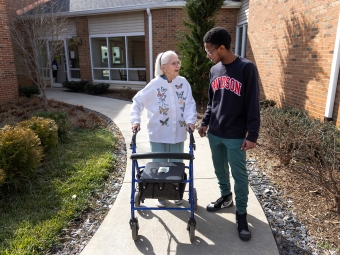 A young man in a black "Davidson" sweatshirt walks along a paved outdoor path and talks with an elderly woman using a blue rolling walker.