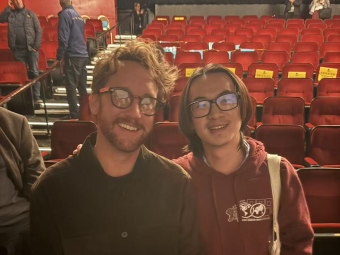 Two men wearing glasses smile while standing together in a theater with rows of red velvet seats in the background.