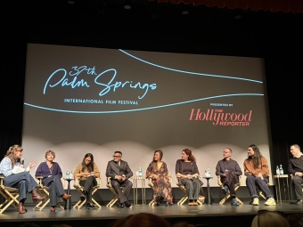 A diverse panel of nine filmmakers sits in director's chairs on a stage in front of a large screen displaying the 37th Palm Springs International Film Festival logo.