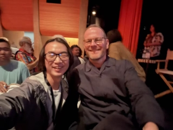A selfie of two men smiling in a dimly lit theater setting with red curtains and wooden paneling in the background.