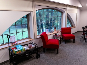 A bright, modern indoor seating area featuring two red armchairs and a small side table positioned in front of large arched windows overlooking a green, wooded landscape.