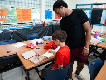 A young man in a black t-shirt leans over to watch a boy in a red shirt drawing in a sketchbook at a classroom desk.
